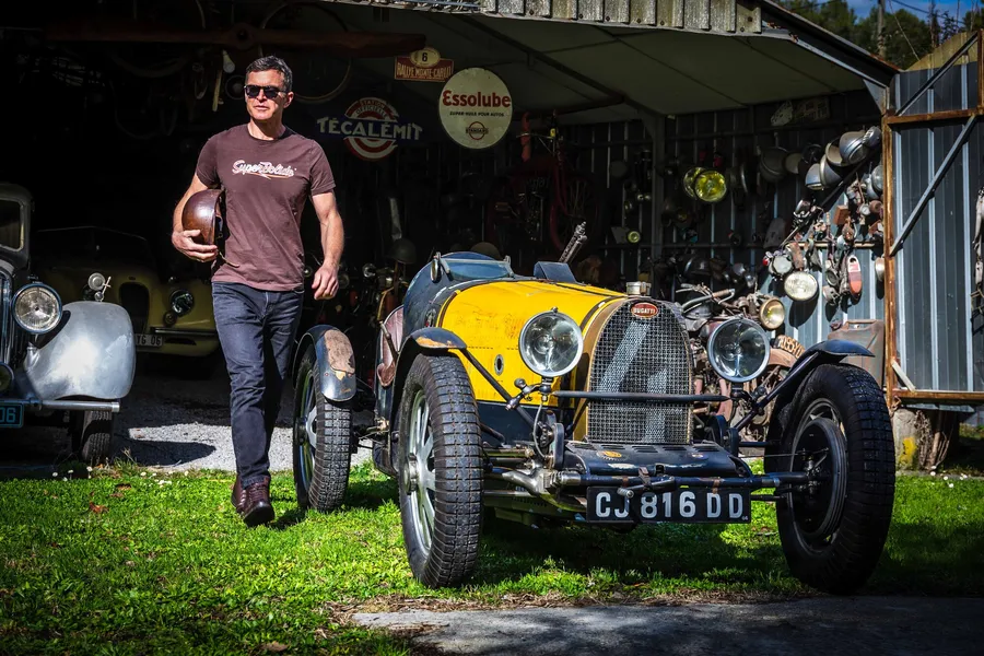 Philippe Lebeau, fondateur de SuperBolide, lunettes de soleil, dans son atelier entouré d'automobiles de collection