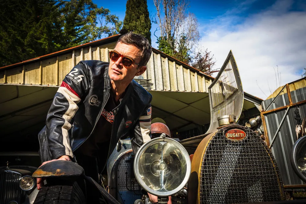 Portrait de Philippe Lebeau, fondateur et pilote de SuperBolide, lunettes de soleil et blouson de course, auprès d'une automobile de collection