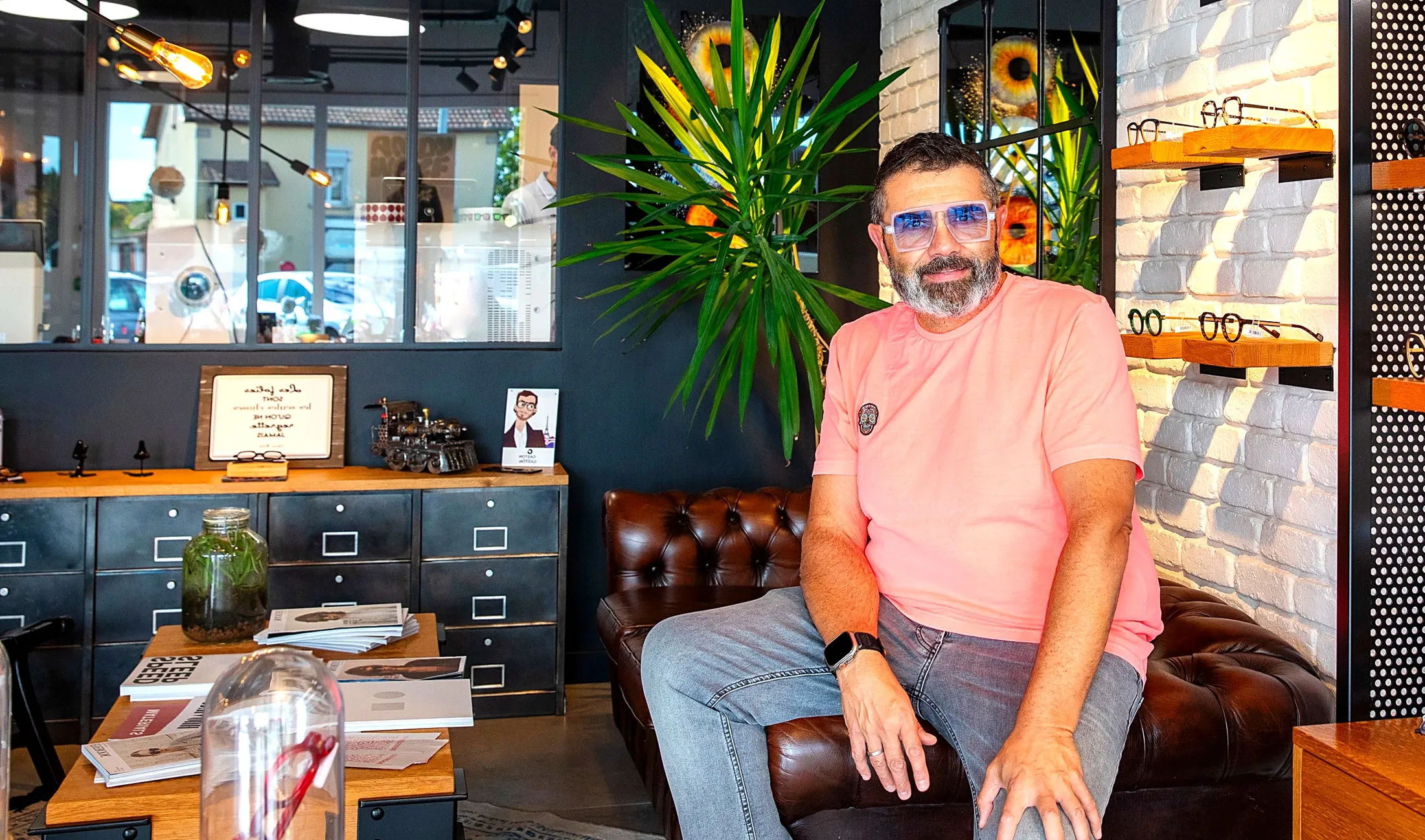 Jérôme, fondateur de La Boutique des Lunetiers, dans le coin salon de la boutique à Essert, montures de créateurs visibles sur l'étagère derrière lui