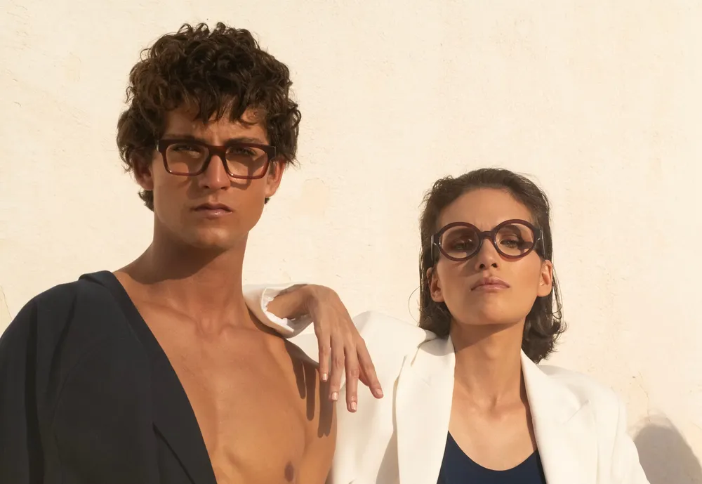 Portrait à contre-jour d'un jeune homme portant une monture optique carrée en acétate transparent Lazuli Eyewear, chemise en lin bleu clair, ciel lumineux en arrière-plan