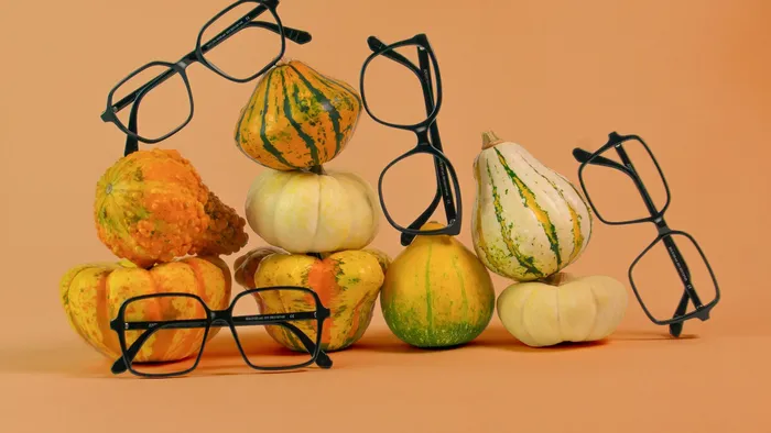 Quatre montures optiques sombres sont disposées parmi un assortiment de courges sur un fond orange.