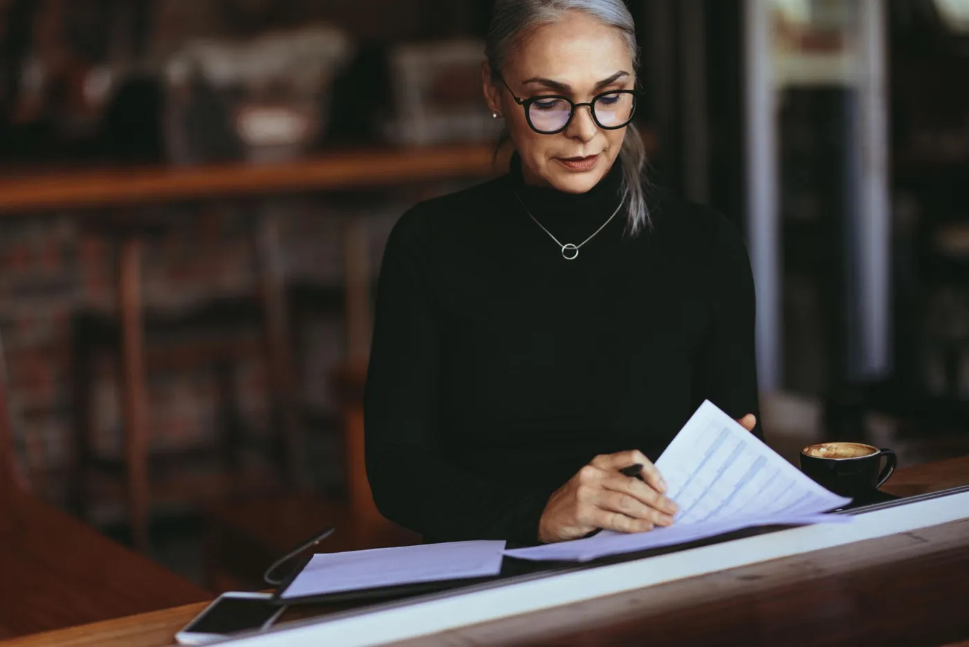 Femme aux cheveux gris portant des lunettes, penchée sur des documents, une tasse de café à côté
